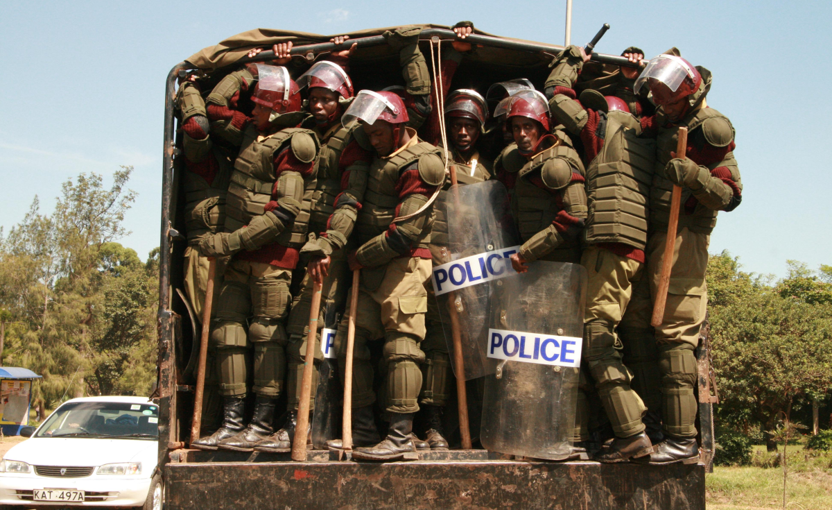 Un grand nombre de policiers armés de clubs attendent de sauter d'une camionnette au Kenya. La police porte beaucoup d'armures, de boucliers, de casques et de matraques.