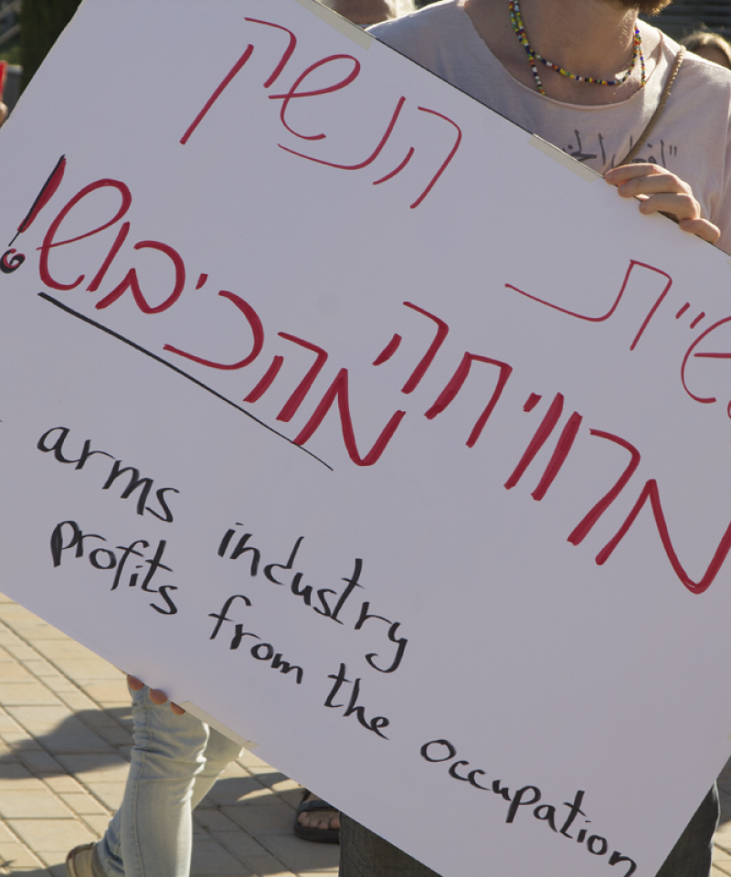 A protester outside the ISDEF arms fair holds a sign reading "The arms industry profits from the occupation"