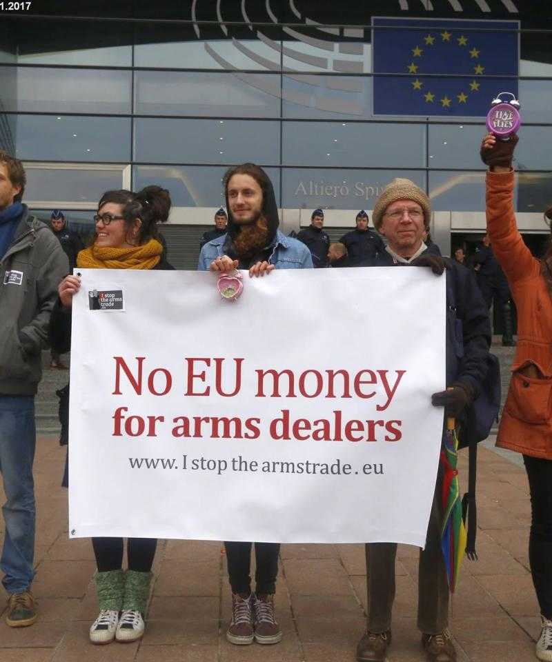 A line of campaigners stand with a banner reading "No EU money for arms dealers", in front of a building flying the EU flag