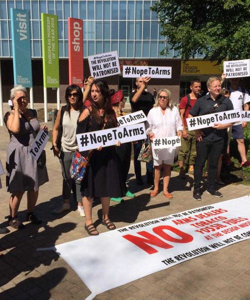A large group of activists stand with placards reading "Nope To Arms" outside the Design Museum in London