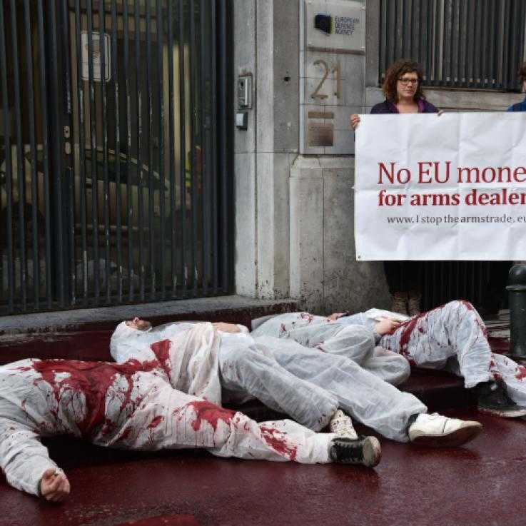 Three activists dressed in white overalls lie in a pool of fake blood. Behind them stand two more activists holding a banner reading "no money for arms dealers"