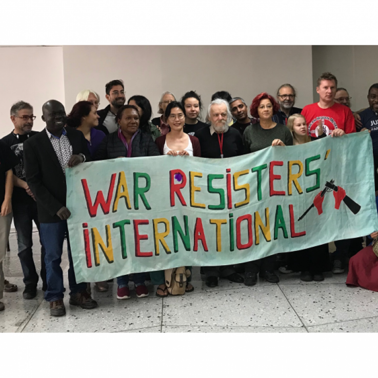 Dos docenas de personas se paran detrás de una pancarta que dice "Internacional de Resistentes a la Guerra"