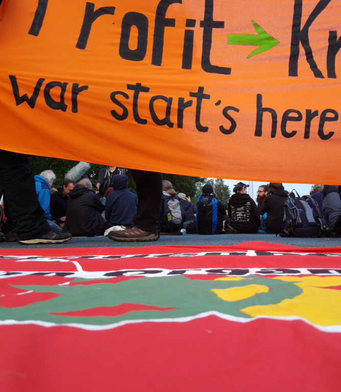Protesters sit below an orange banner reading "war starts here"