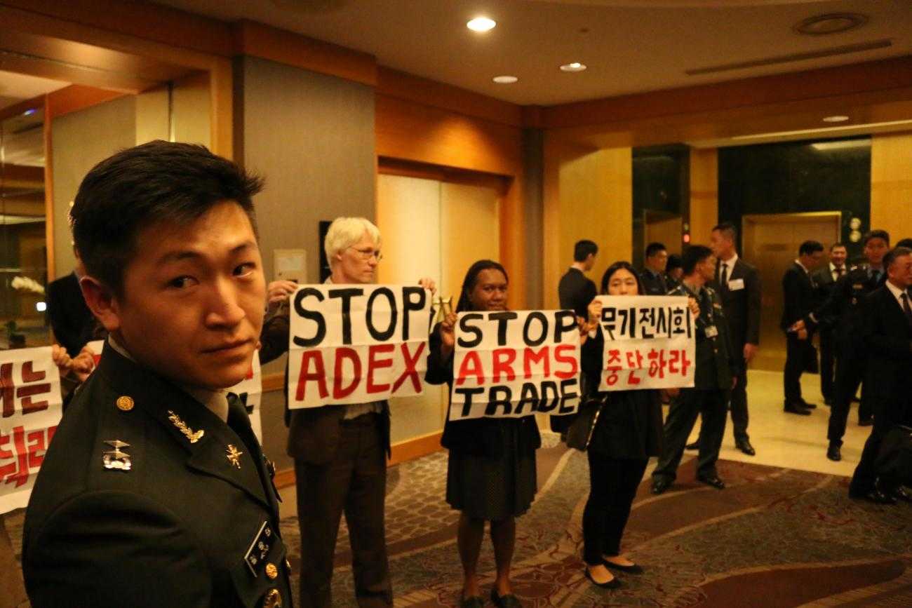International activists inside the arms dealers welcome dinner at ADEX 2015 in Seoul. Protesters hold up banners saying 'Stop ADEX' whilst a soldier looks on