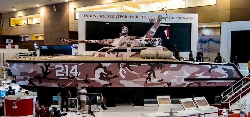 CMI Defences "tank boat" - a catamaran with a large turret, exhibited indoors at a trade show. The boat is painted in brown camoflage patterns.
