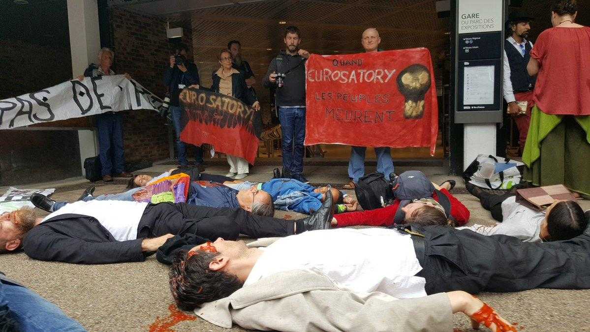 A large group of people lie on the floor as part of a "die-in". In the background other protesters stand with banners.