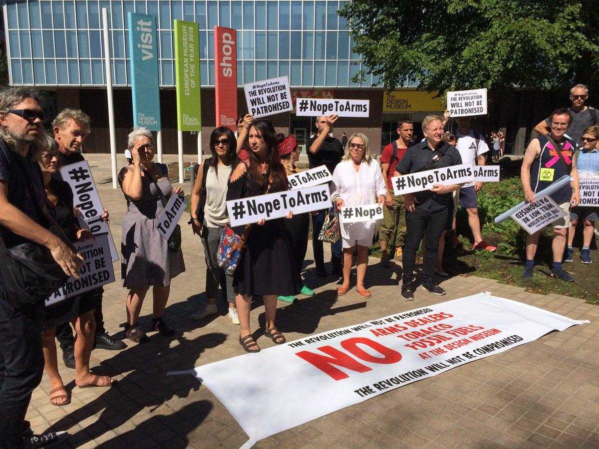 A large group of activists stand with placards reading "Nope To Arms" outside the Design Museum in London