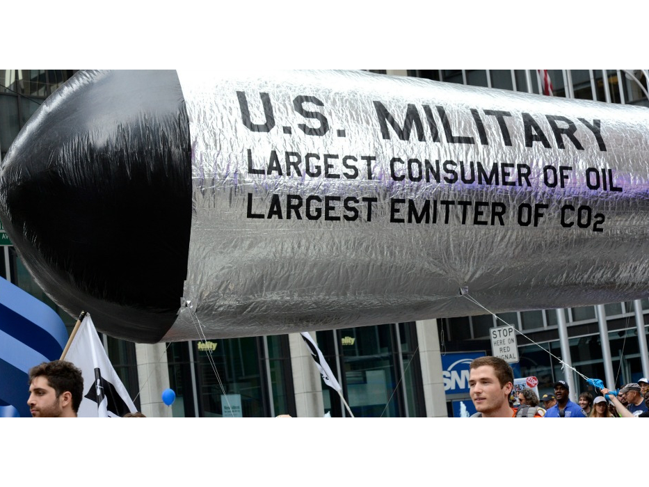 A number of people walk with a large inflatable missile reading "US military: largest user of oil and largest emitter of Co2"