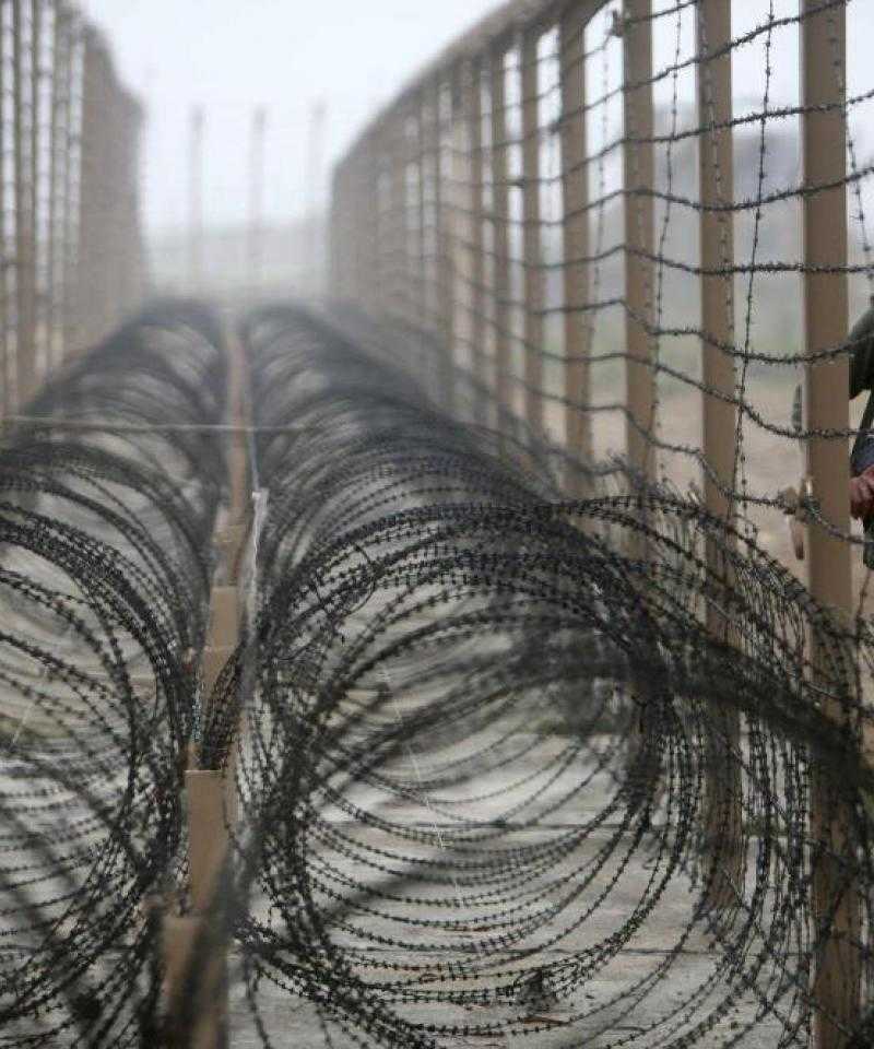 Coils of barbed wire lie between two fences with a soldier patrolling the other side of the right-hand fence