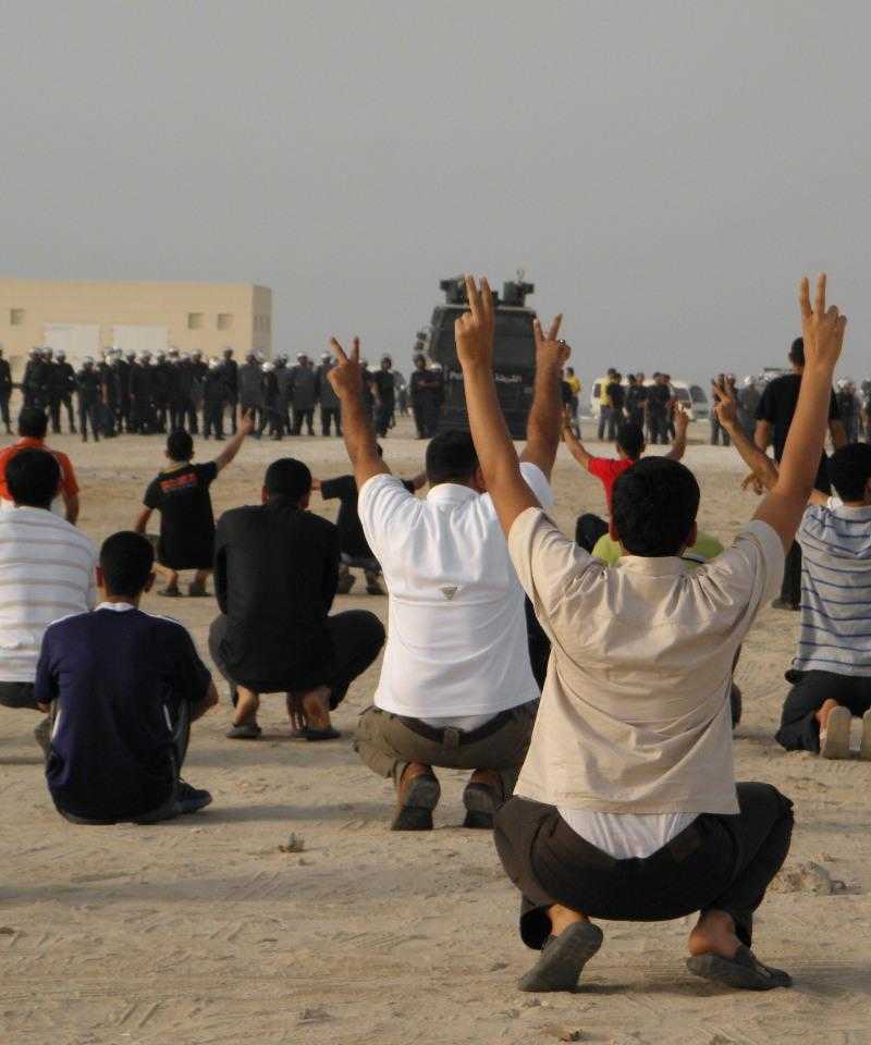 Protesters squat in the foreground with their arms in the air making peace signs, their backs to the camera.  Facing them is a line of police dressed in riot gear with a large, armoured vehicle in the middle of the line.