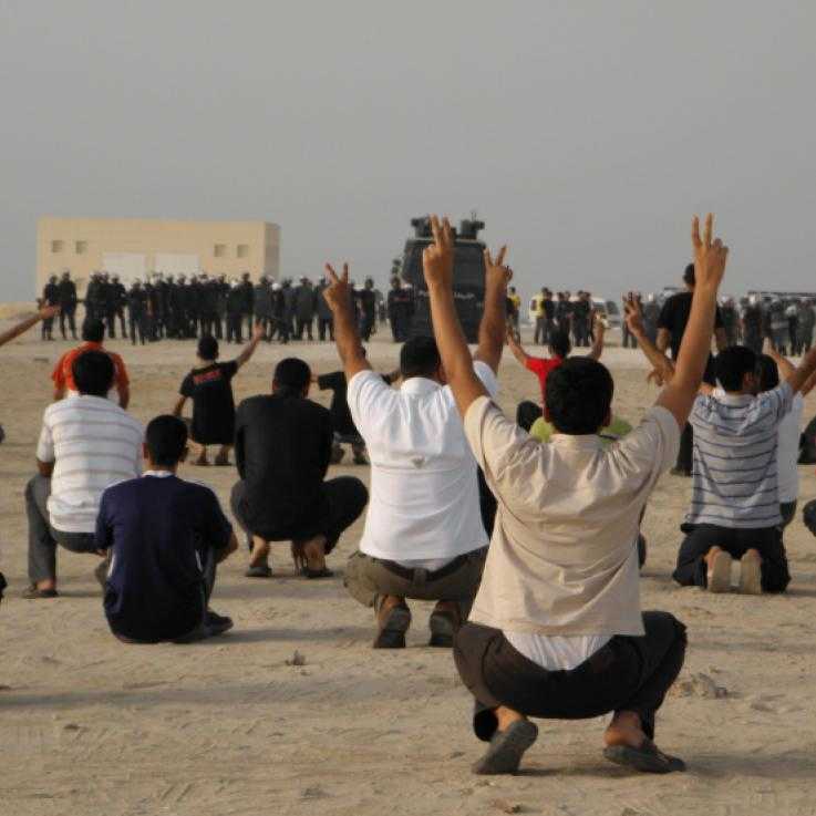 Protesters squat in the foreground with their arms in the air making peace signs, their backs to the camera.  Facing them is a line of police dressed in riot gear with a large, armoured vehicle in the middle of the line.