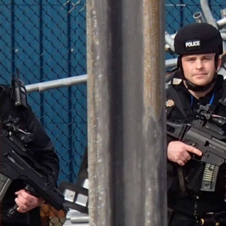 Two members of the heavily armed Civil Nuclear Constabulary stand behind a fence. They are wearing helmets, bullet proof vests, and carrying large guns.