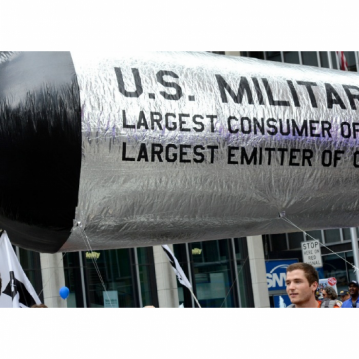 Un número de personas camina con un gran misil inflable que dice "Ejército de EE. UU.: El mayor usuario de petróleo y el mayor emisor de CO2"