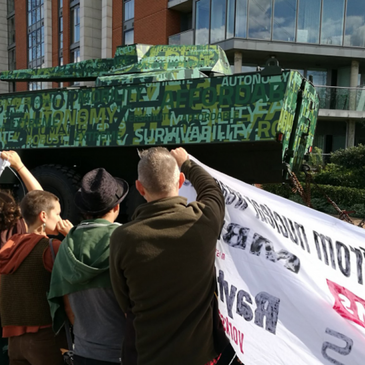 A tank entering the DSEI arms fair, covered in marketing slogans. In front stand a crowd holding banners
